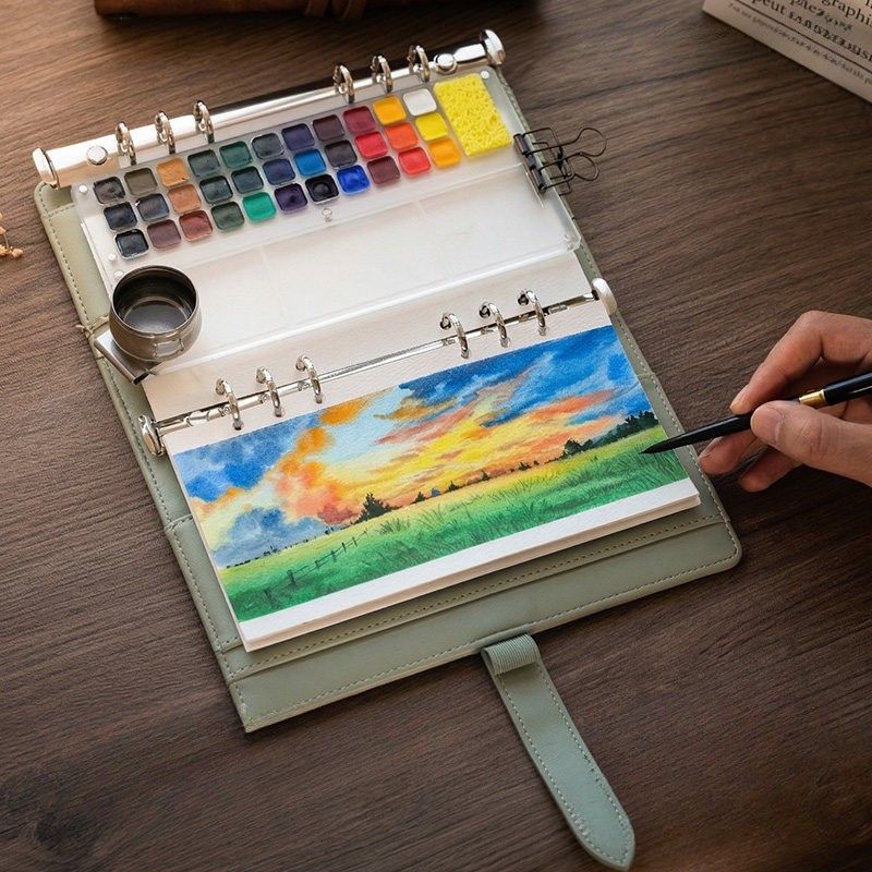 Open Spruce Green Voyager kit on a wooden table with its 33-pan magnetic palette, absorbent sponge and water pot, painting a sunset meadow across the A5 page