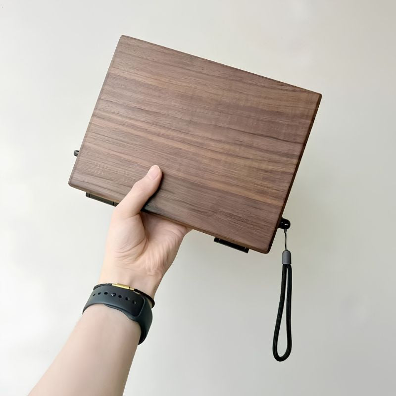 Hand holding the closed walnut Master Edition box from above, showing the wood-grain lid and black wrist strap
