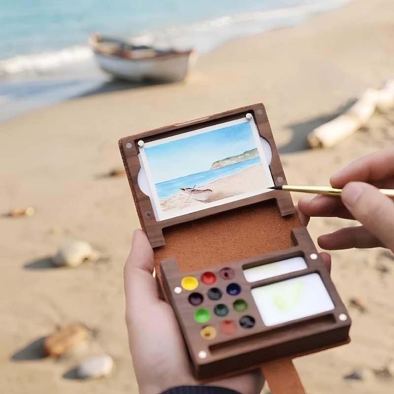 Hands holding the open walnut folio kit on a beach, painting a boat scene with 12 watercolor pans