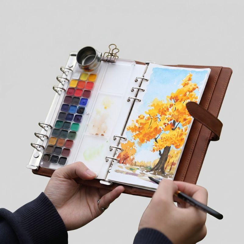 Hands holding the open ArtMate Voyager leather watercolor kit, painting an autumn tree beside the 33-pan palette, water pot and binder clip