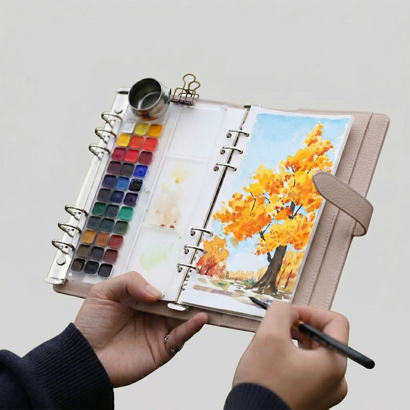 Hands holding the open Cream Apricot Voyager watercolor kit, painting an autumn tree with the 33-pan palette and water pot