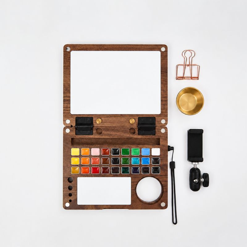 Flat lay of the open walnut Master Edition kit — 27-pan palette, mixing tile and water-pot well — beside a brass water pot, rose-gold binder clip, wrist strap and phone-holder clamp